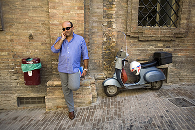 Italy - Urbino - A man talks on his mobile telephone while leaning against a wall next to his Vespa